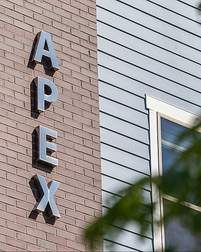 Modern exterior of APEX building with sleek signage and large windows framed by lush green leaves.