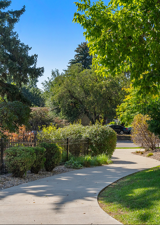 Charming apartment exterior with lush greenery, mature trees, and a welcoming pathway under a bright blue sky.