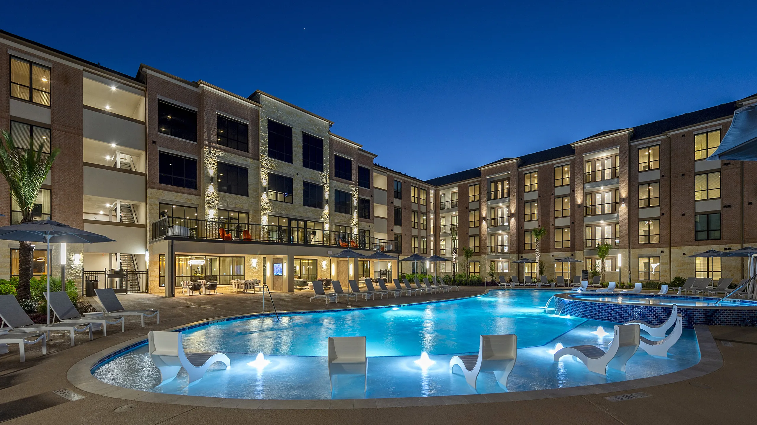 Illuminated apartment complex at dusk featuring a sparkling swimming pool with modern lounge chairs.