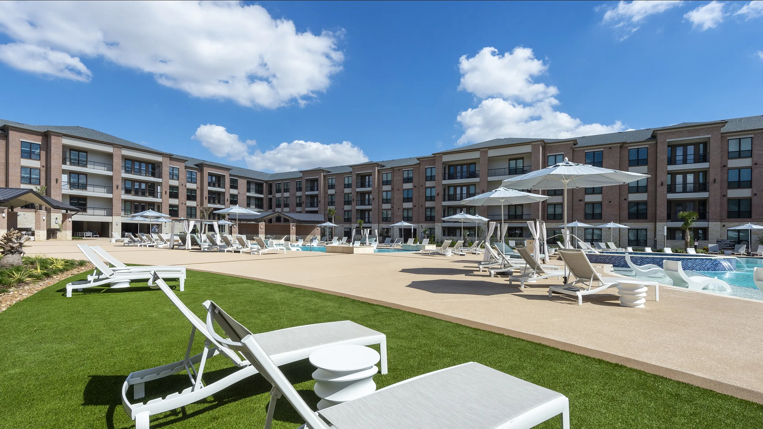 Elegant apartment complex with a sprawling outdoor pool area, surrounded by sun loungers and umbrellas.