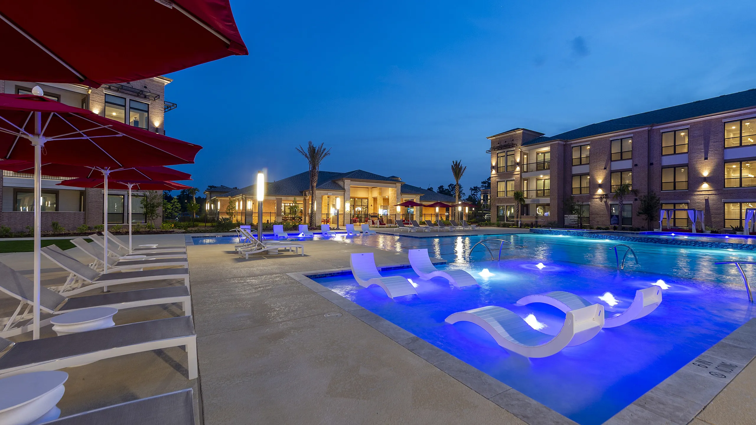 Luxurious apartment complex at dusk with a stunning illuminated swimming pool, comfortable lounge chairs, and elegant red umbrellas, offering a serene and upscale living environment.