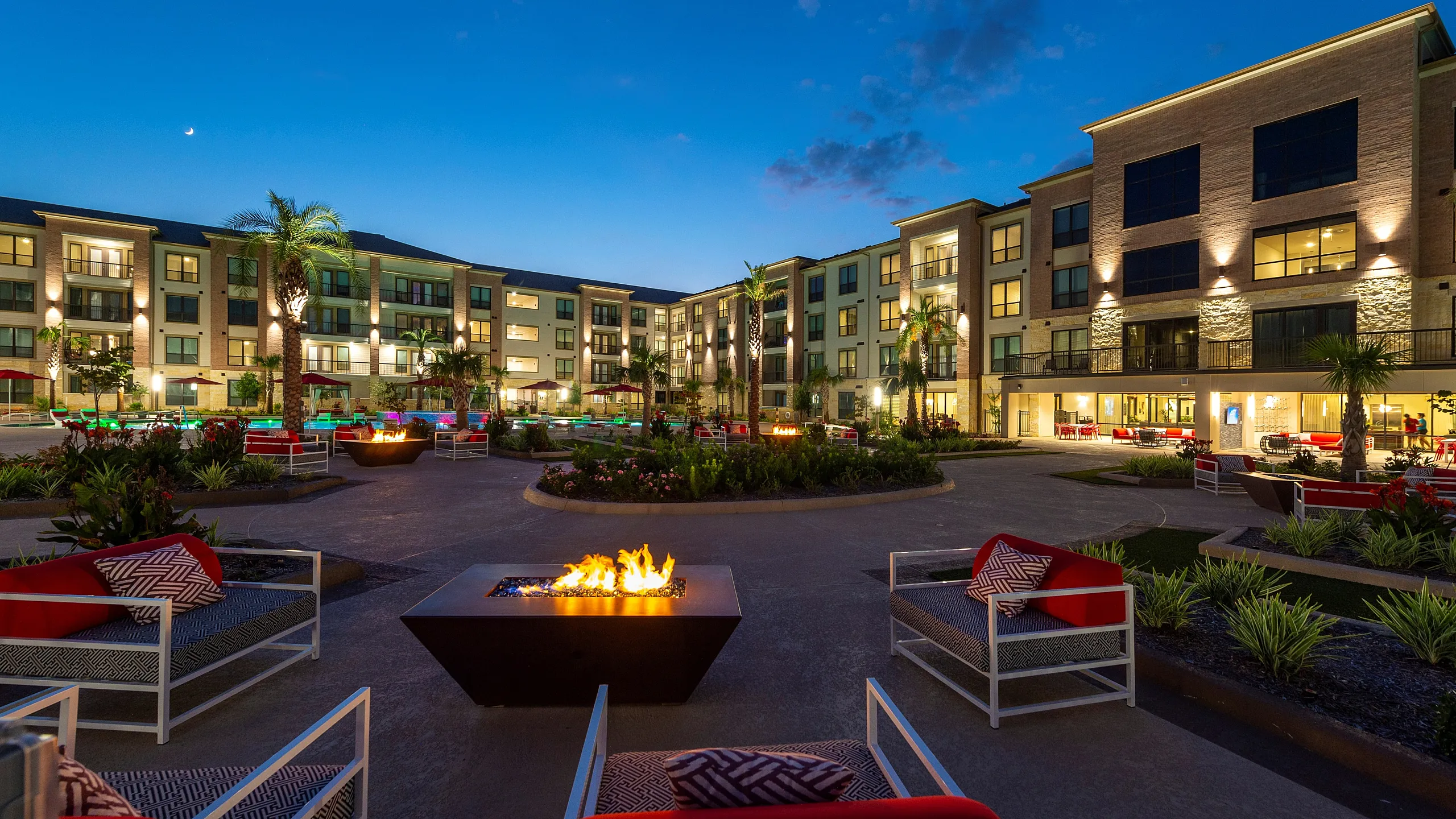 Luxurious apartment complex at dusk featuring modern architecture, illuminated pathways, cozy fire pits, and lush landscaping.