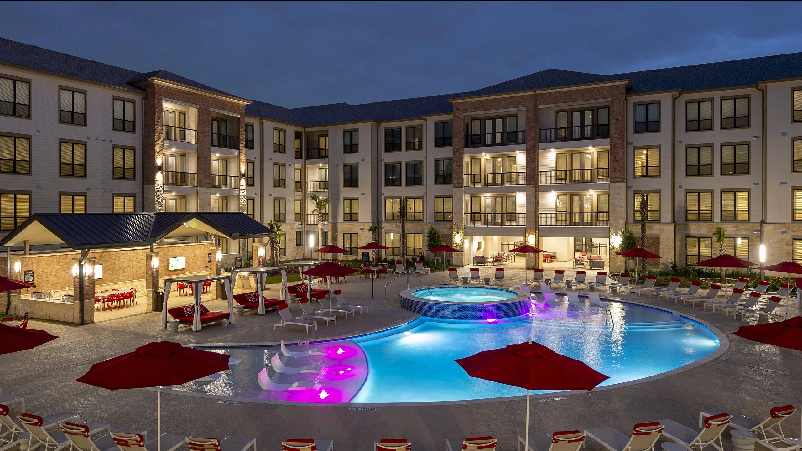 Luxurious apartment complex with an illuminated pool, stylish lounge chairs, and modern cabanas, creating a resort-like atmosphere.