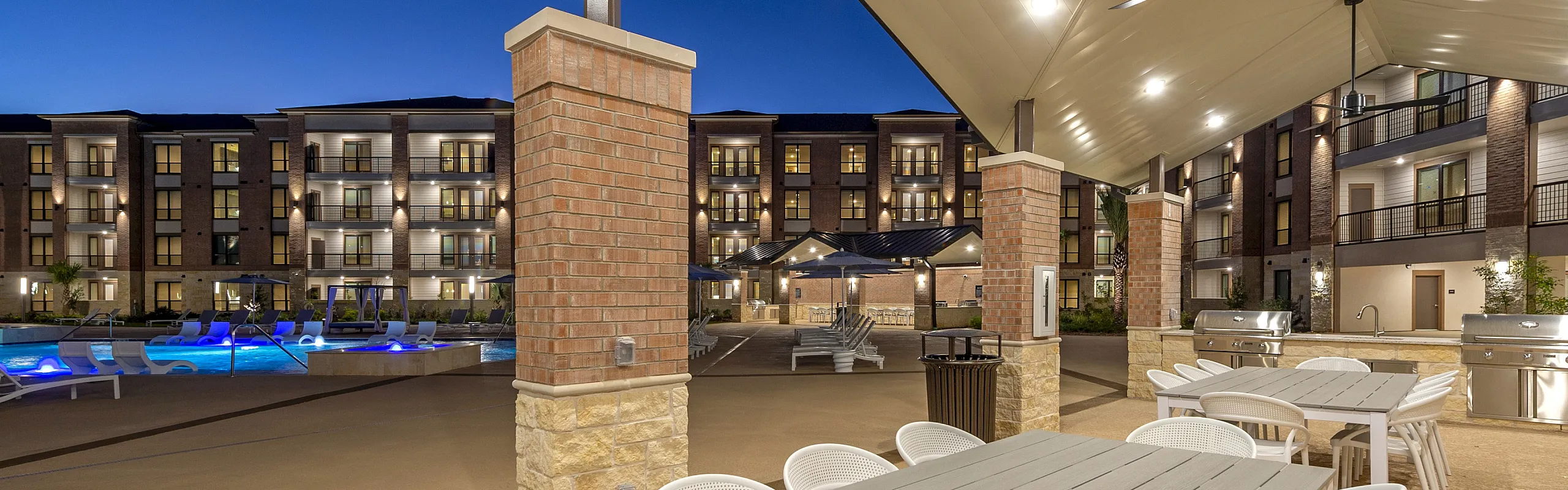 Elegant outdoor dining area under a modern pavilion with views of a sparkling pool and luxurious apartment complex at twilight.