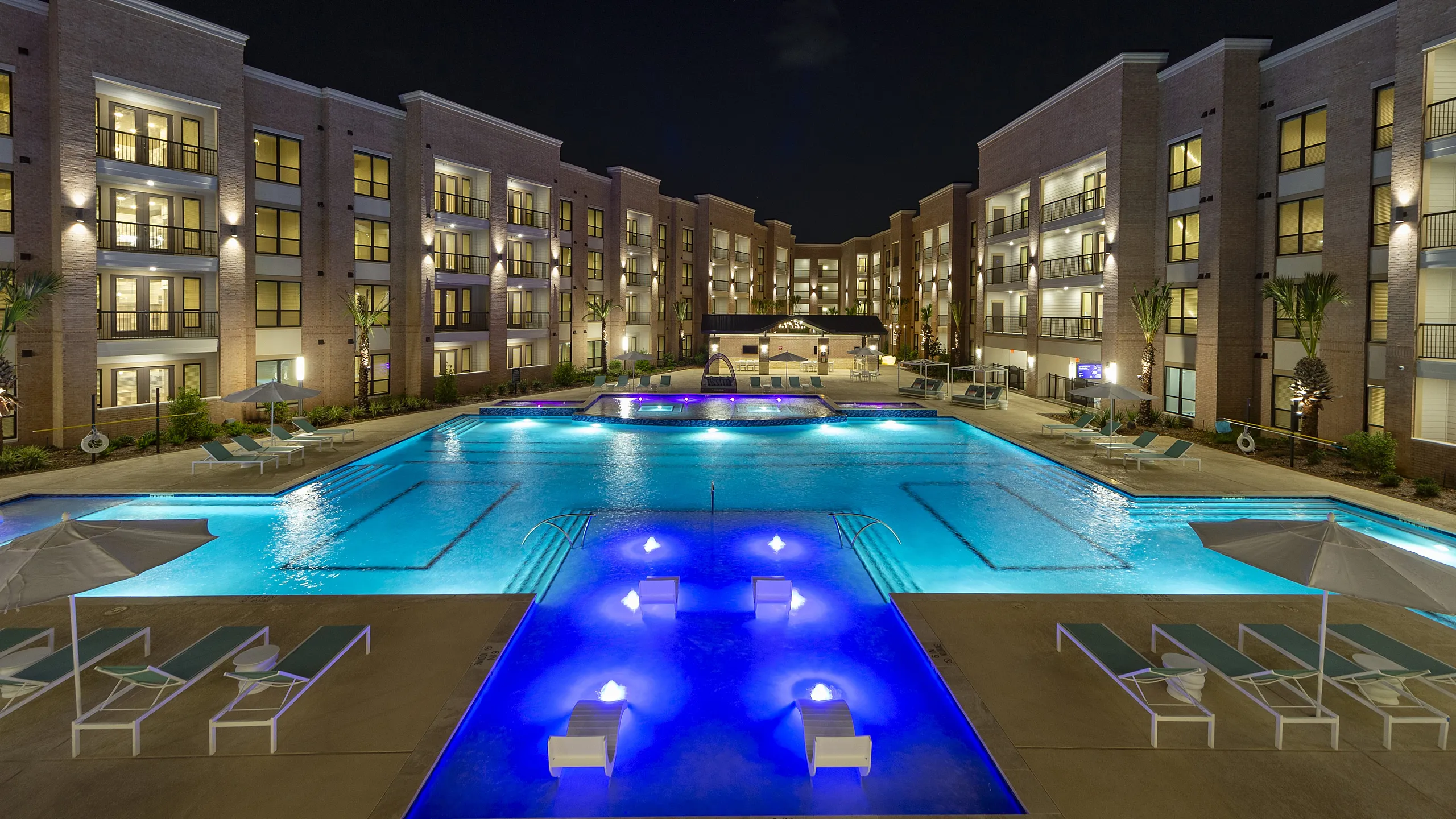 Stunning nighttime view of a luxury apartment complex with an illuminated swimming pool and elegant poolside seating.
