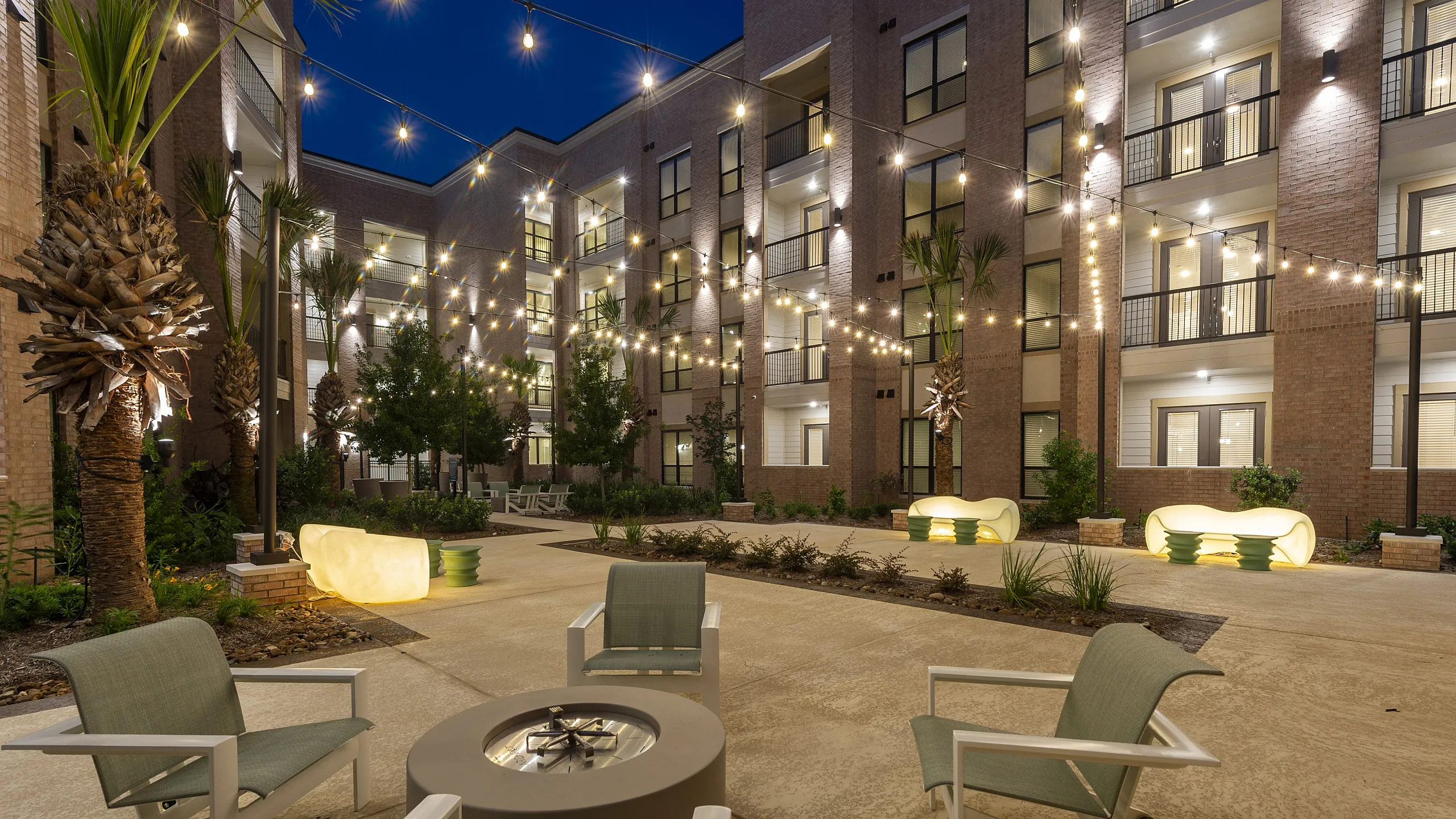 Charming apartment courtyard at night, illuminated by string lights, featuring modern seating and lush landscaping.