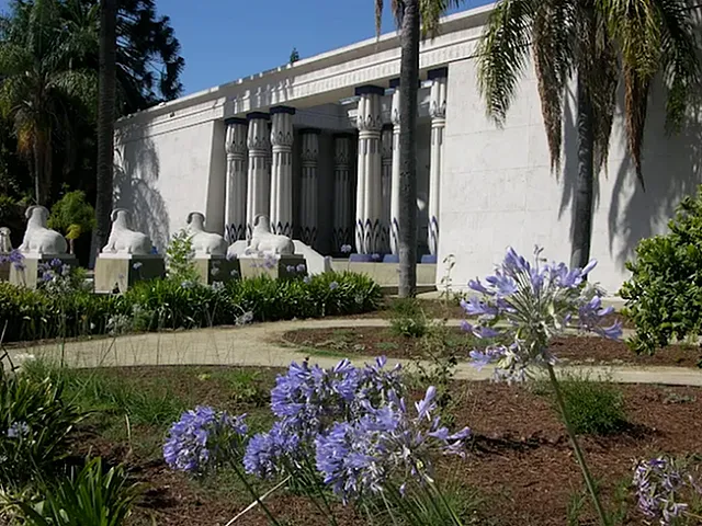 Elegant white building with majestic columns and lush landscaping, surrounded by vibrant purple flowers and palm trees for a serene ambiance.