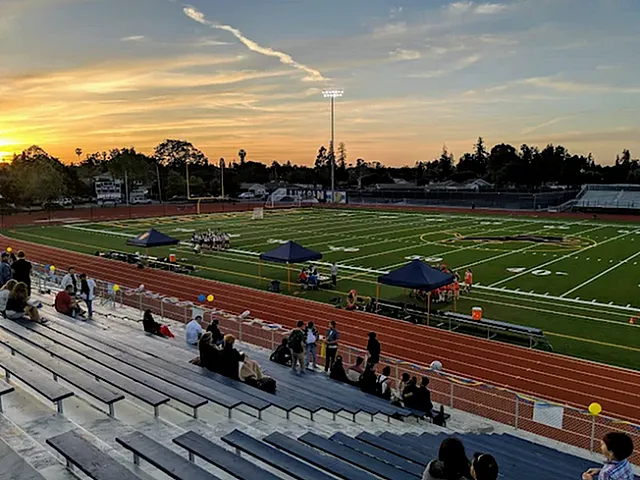 Vibrant sports field at sunset with spectators on bleachers, featuring well-maintained green turf, surrounded by a red running track.