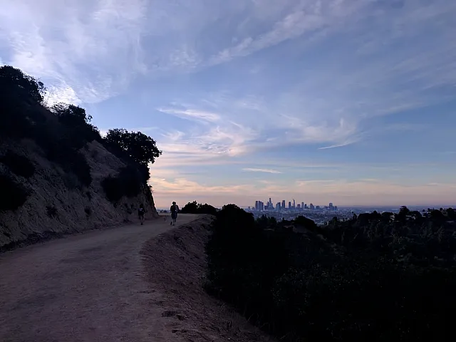 Scenic hillside path with people walking, offering stunning views of a city skyline at dusk.