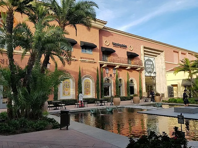 Charming shopping plaza with lush palm trees, elegant architecture, and a tranquil reflecting pool.