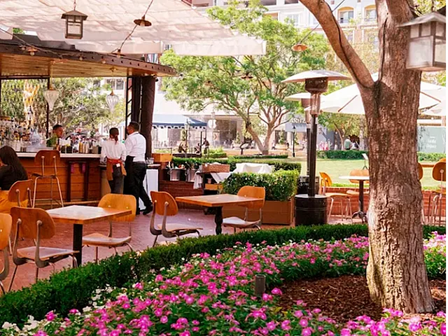 Charming outdoor cafe with colorful flower beds, shaded seating, and lush tree canopy, perfect for a relaxing meal.