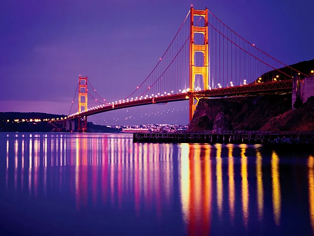 Enchanting view of a glowing suspension bridge at twilight, reflecting vibrant colors on calm waters.