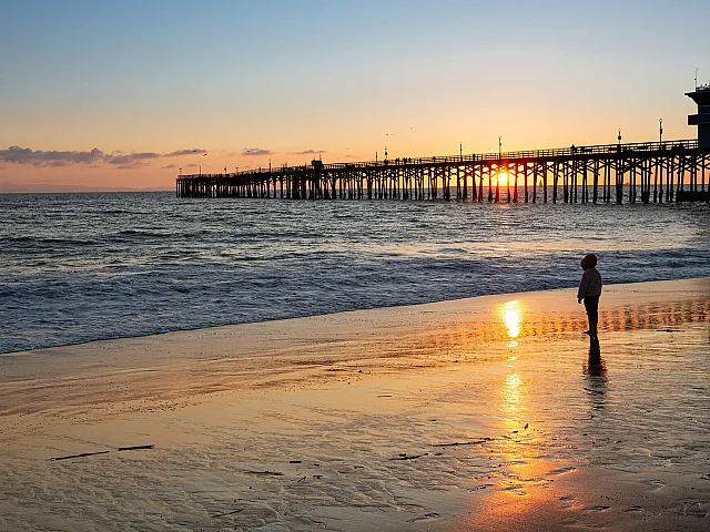 Breathtaking view of a serene beach at sunset with a silhouette of a child and a long wooden pier under a vibrant sky.