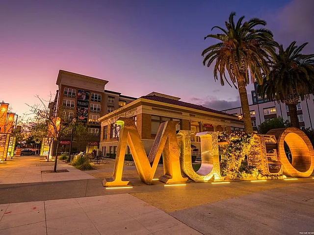 Charming urban evening scene featuring illuminated 'Muzeo' sign, surrounded by palm trees and modern architecture under a picturesque dusk sky.