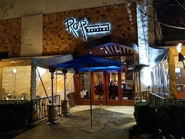 Inviting restaurant entrance with warm lighting, outdoor seating under blue umbrellas, and a charming wooden facade.