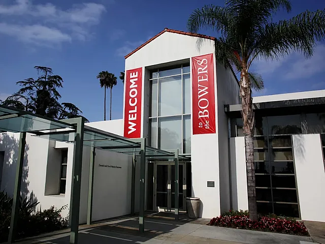 Modern museum entrance with striking glass canopy and palm trees under a clear blue sky.