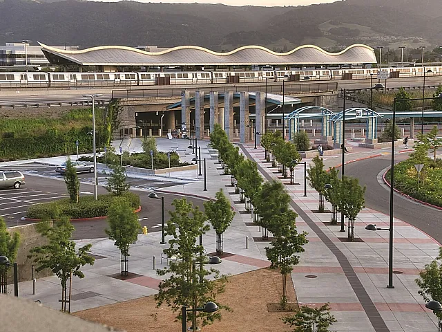 Scenic view of a modern train station surrounded by lush greenery and a well-maintained plaza, set against a backdrop of rolling hills.