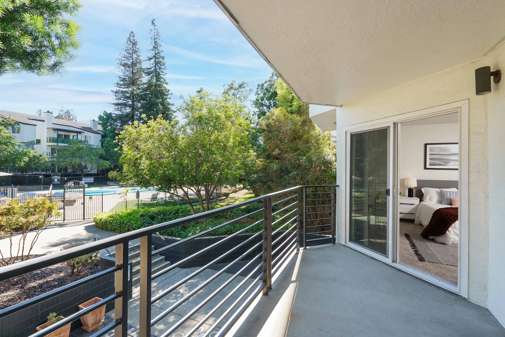 Charming balcony overlooking a lush garden and pool area, with sliding doors leading to a cozy bedroom.