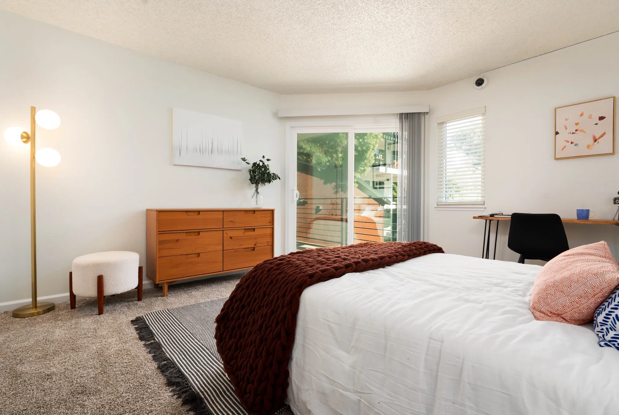 Modern bedroom featuring a cozy bed with decorative pillows, a wooden dresser, floor lamp, and sliding doors leading to a balcony with lush greenery.
