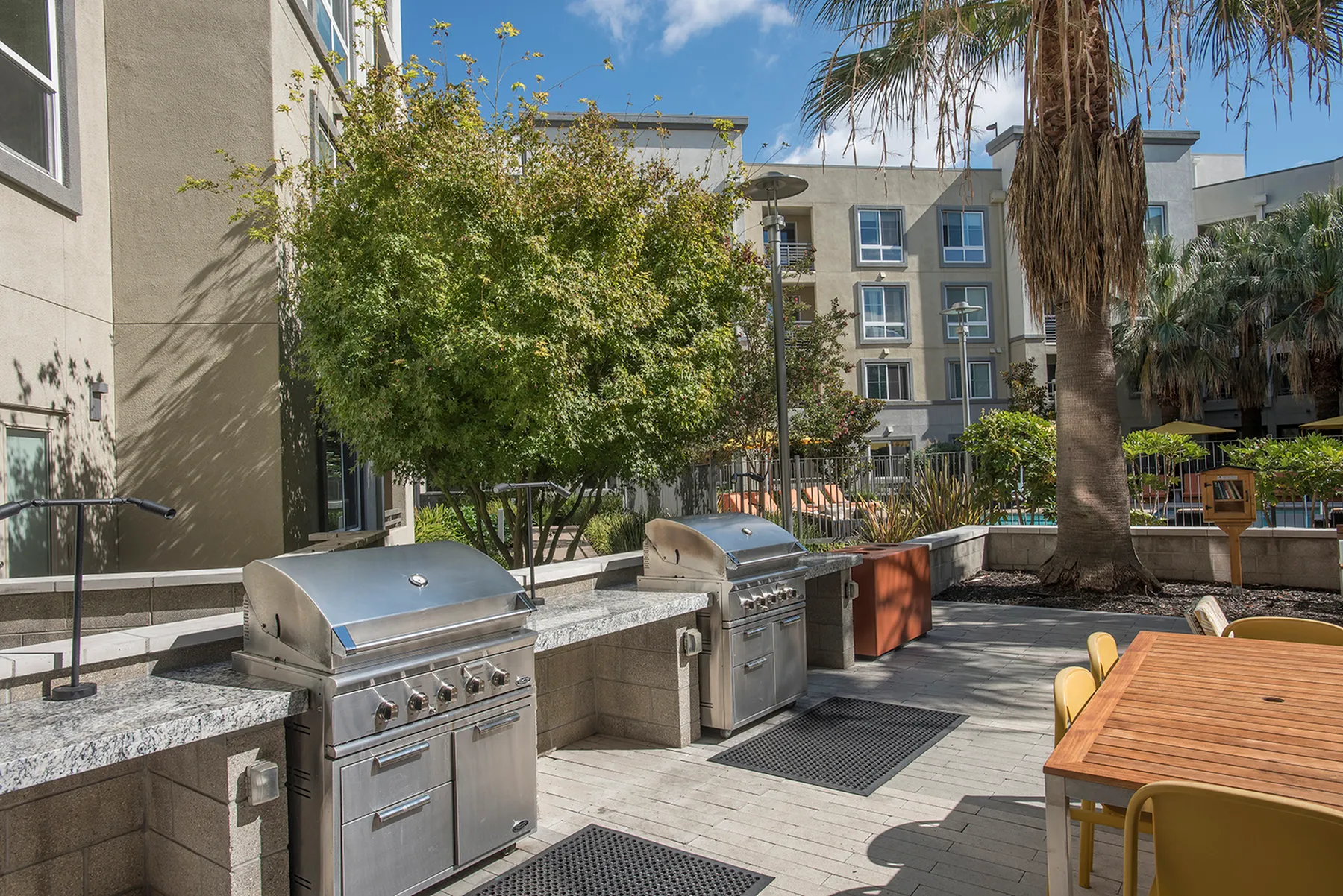 Chic apartment outdoor space with modern stainless steel grills, lush greenery, and a dining area under sunny skies.