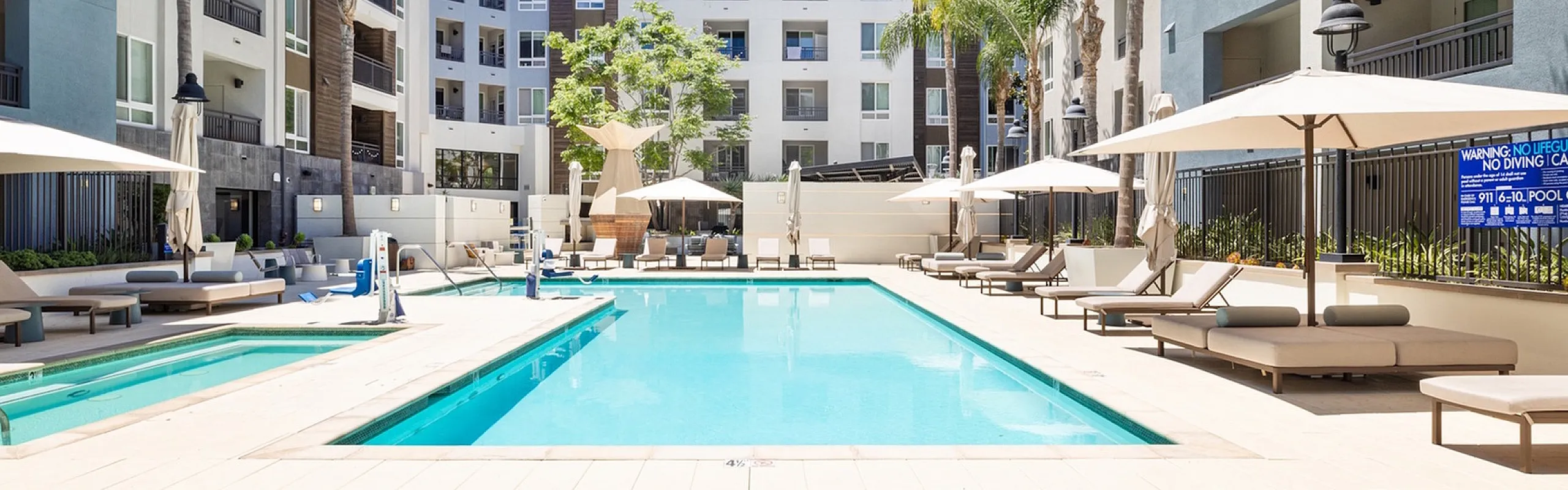 Luxurious apartment complex featuring a sparkling pool surrounded by stylish loungers and vibrant palm trees, offering a perfect retreat in a sunny courtyard.