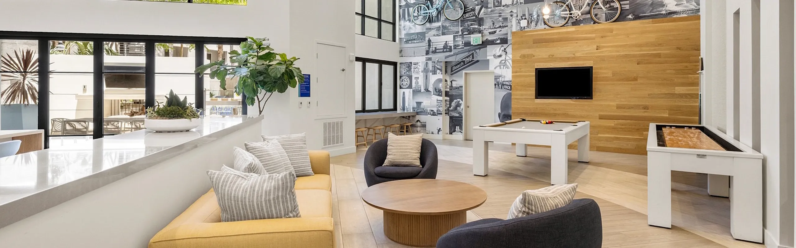 Bright and inviting community lounge with modern decor, featuring a pool table, cozy seating, and large windows overlooking lush greenery.