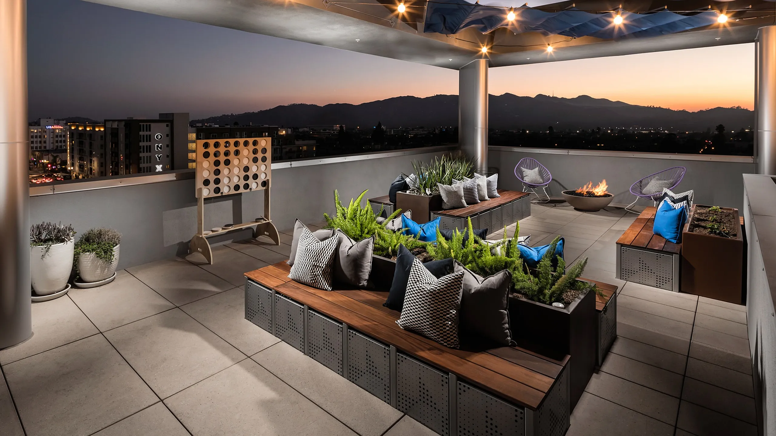 Luxurious rooftop patio with cozy seating, lush greenery, and a fire pit, offering stunning mountain and city views at sunset.