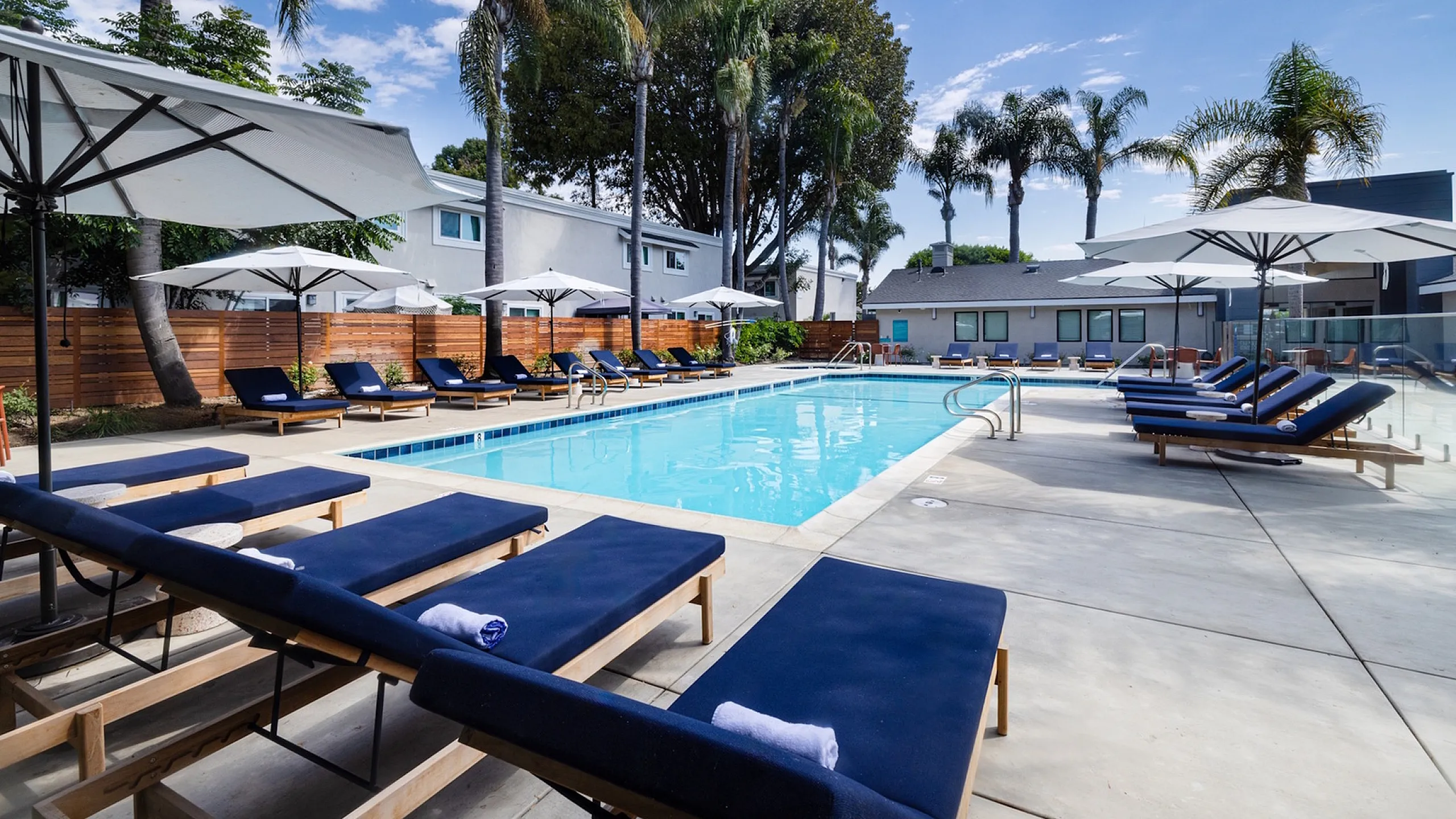 Inviting outdoor pool area with luxurious sun loungers and large umbrellas, surrounded by lush palm trees and modern apartments.