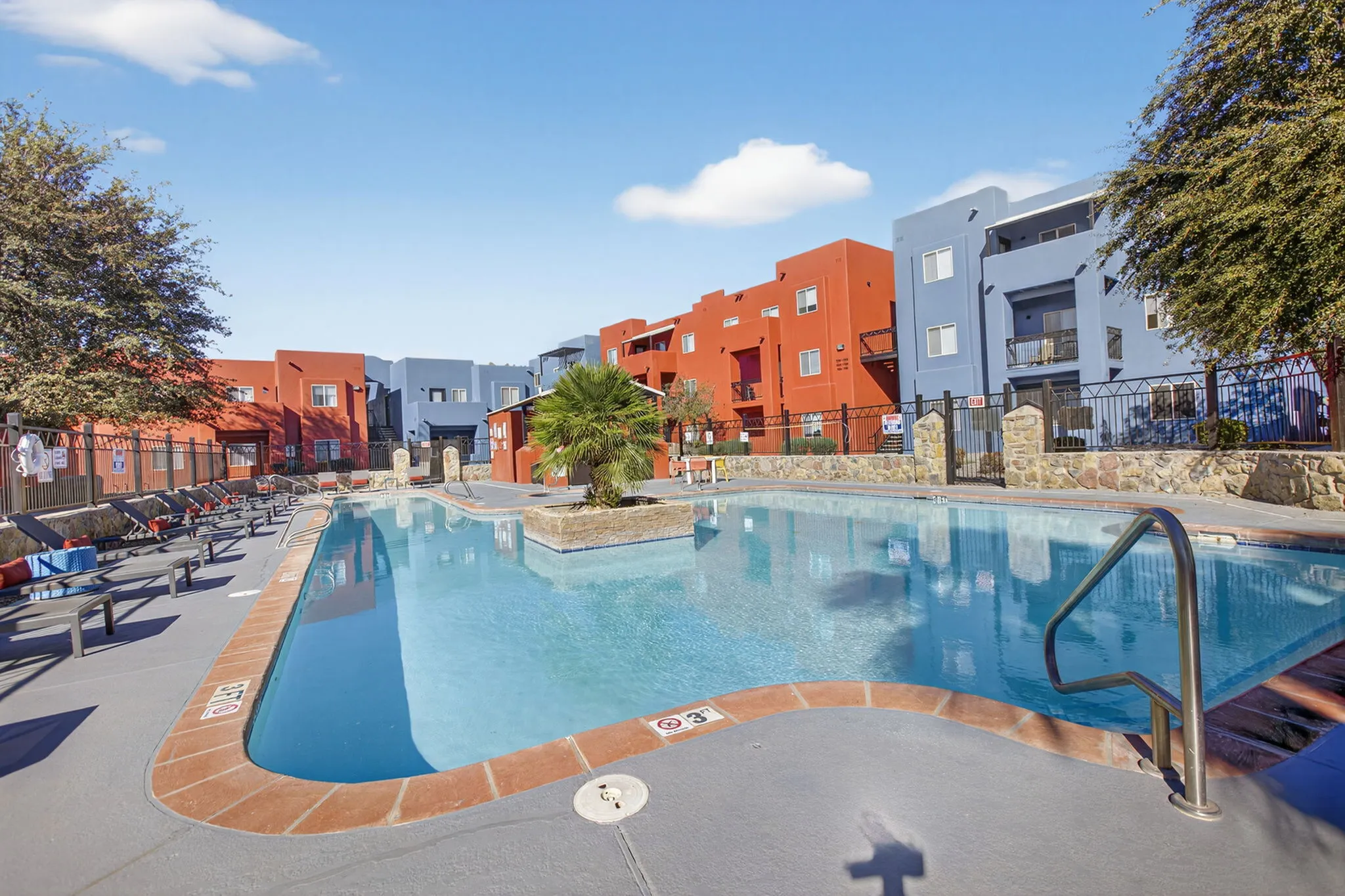 Vibrant apartment complex with red and blue facades featuring a dazzling pool surrounded by modern loungers and lush greenery.