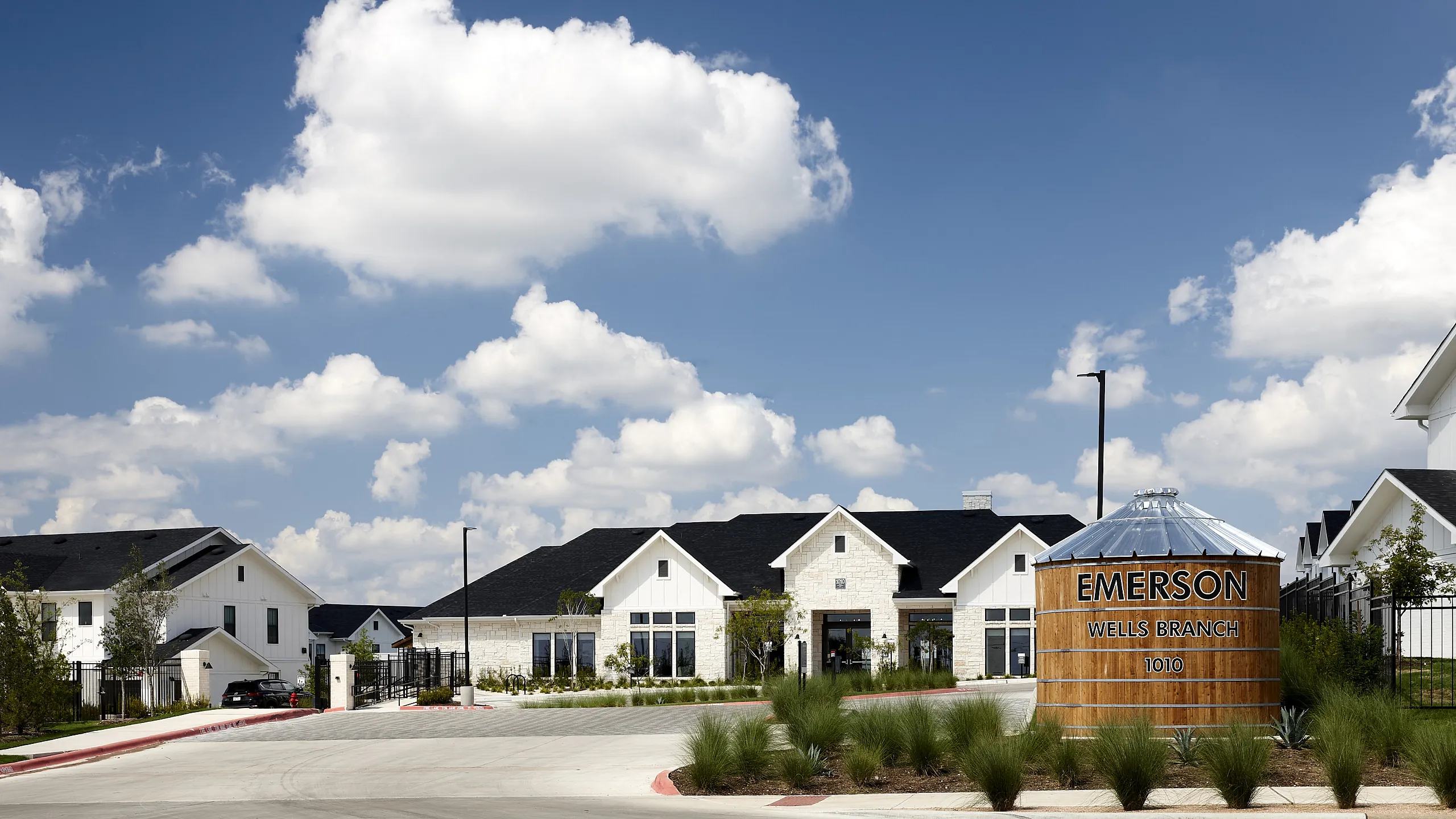 Charming community entrance of Emerson Wells Branch with modern farmhouse architecture and a welcoming sign under a bright blue sky.