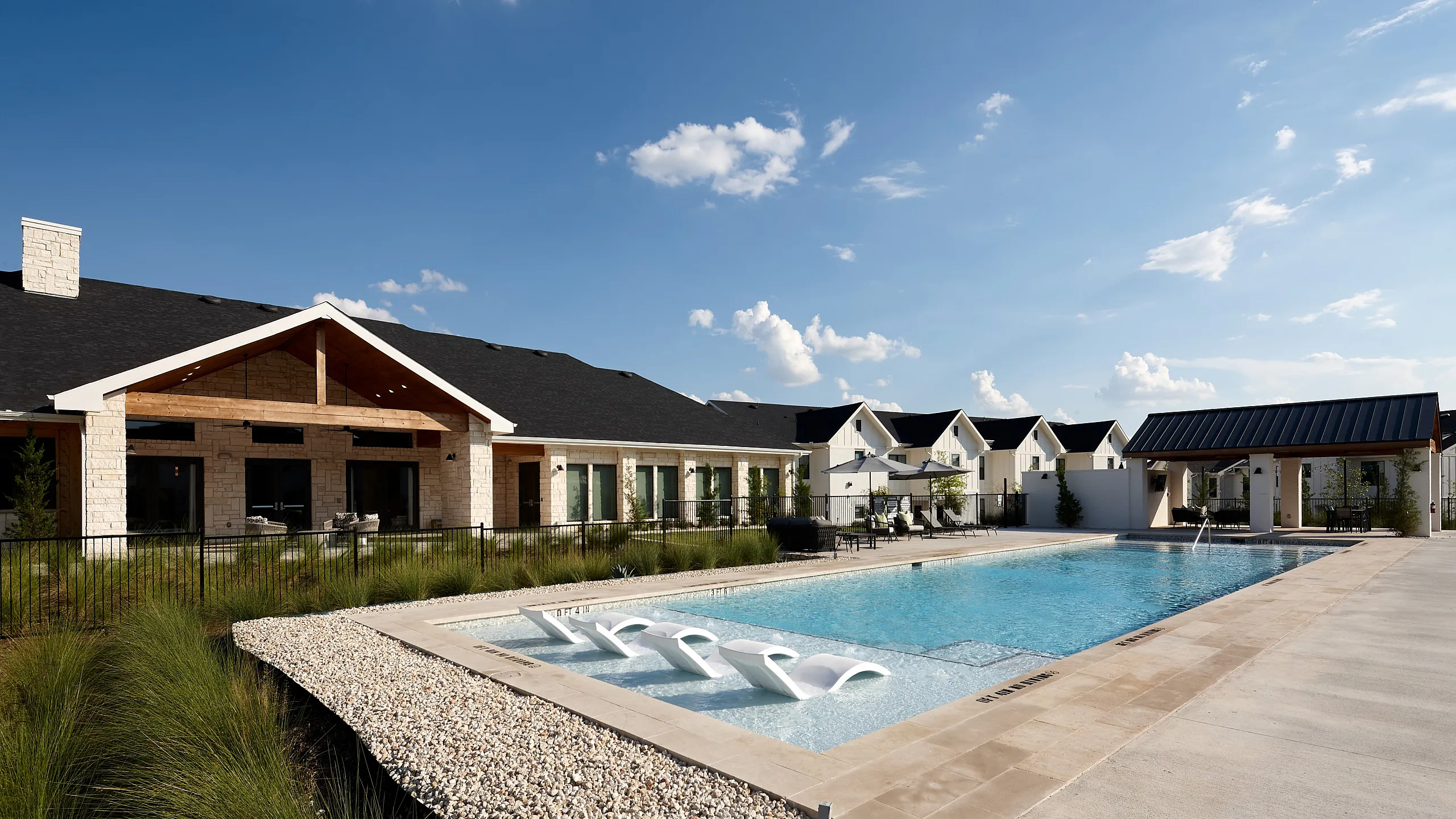 Luxurious outdoor pool area with elegant loungers and a modern clubhouse, under a bright blue sky at a serene residential community.