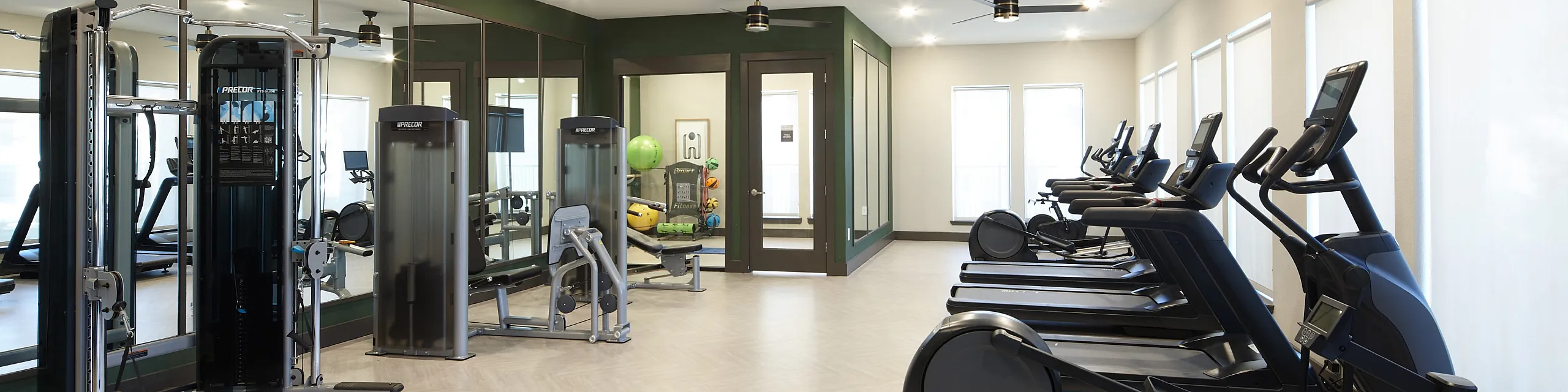 Modern fitness center with sleek exercise equipment including treadmills, rowing machine, and strength training stations. Bright and spacious with large mirrors and ceiling fans for comfort.