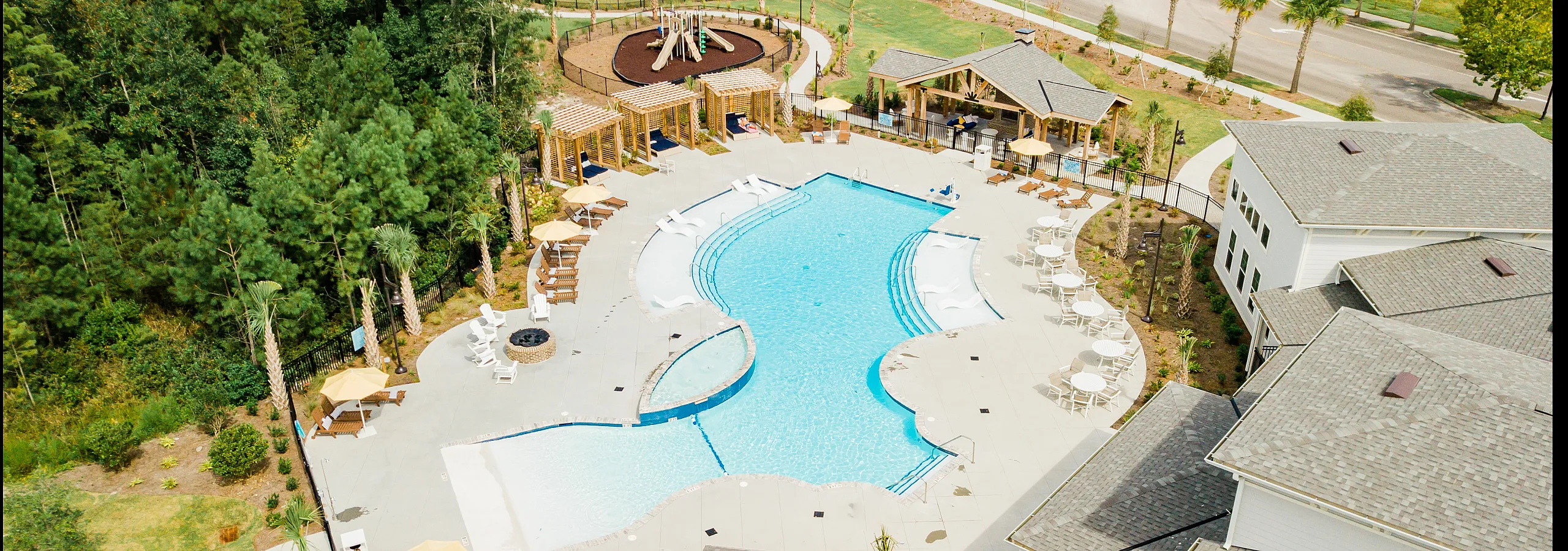 Aerial view of a luxurious apartment complex showcasing a sparkling blue swimming pool, surrounded by stylish sun loungers and umbrellas. The vibrant green landscape and nearby playground create an inviting atmosphere for relaxation and fam