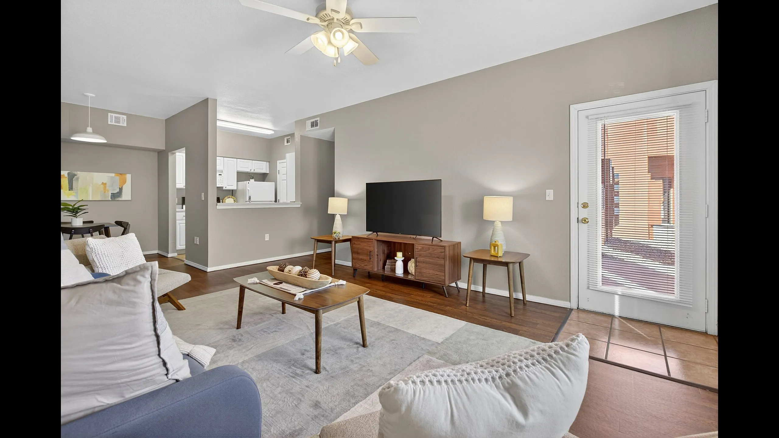 Bright and inviting living room with modern decor, wood flooring, and natural light streaming through glass doors. Open layout with a view into a cozy kitchen.