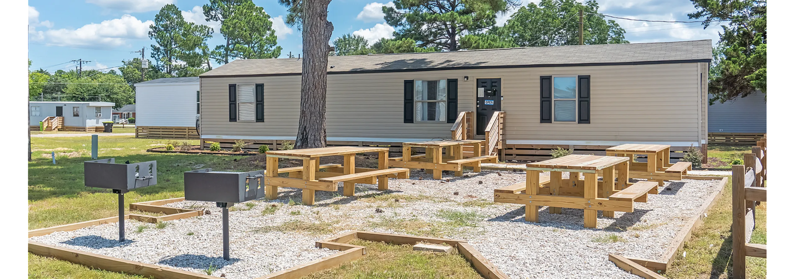 Charming modular home with a welcoming front porch, nestled among lush trees, featuring outdoor picnic areas and grills for community enjoyment.