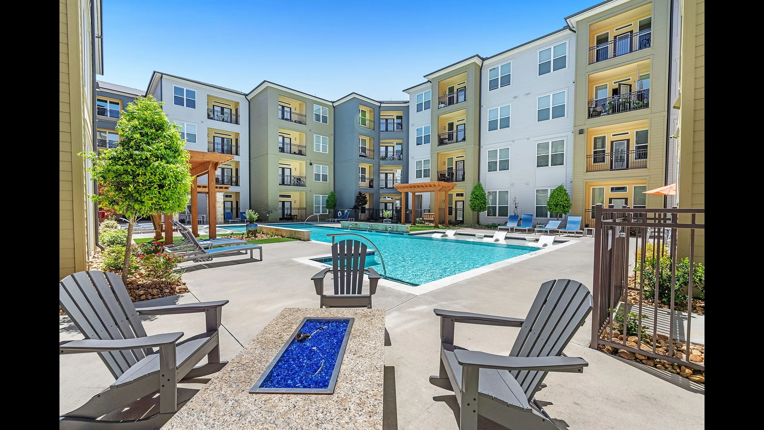 Inviting outdoor area featuring a modern apartment complex with a sparkling pool, sun loungers, and cozy seating under clear blue skies.
