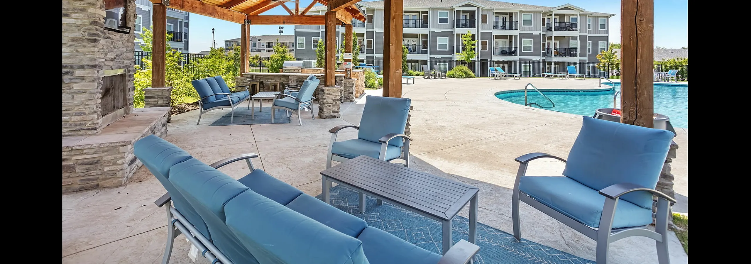 Charming outdoor lounge with cozy seating under a wooden pergola, overlooking a sparkling pool and modern apartment building.