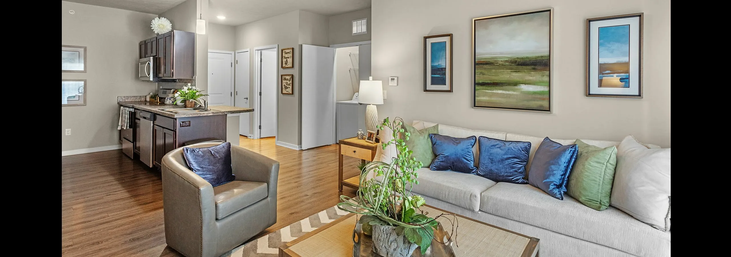 Inviting open-concept living room with stylish decor, ceiling fan, and a modern kitchen, featuring warm wood floors and elegant artwork.