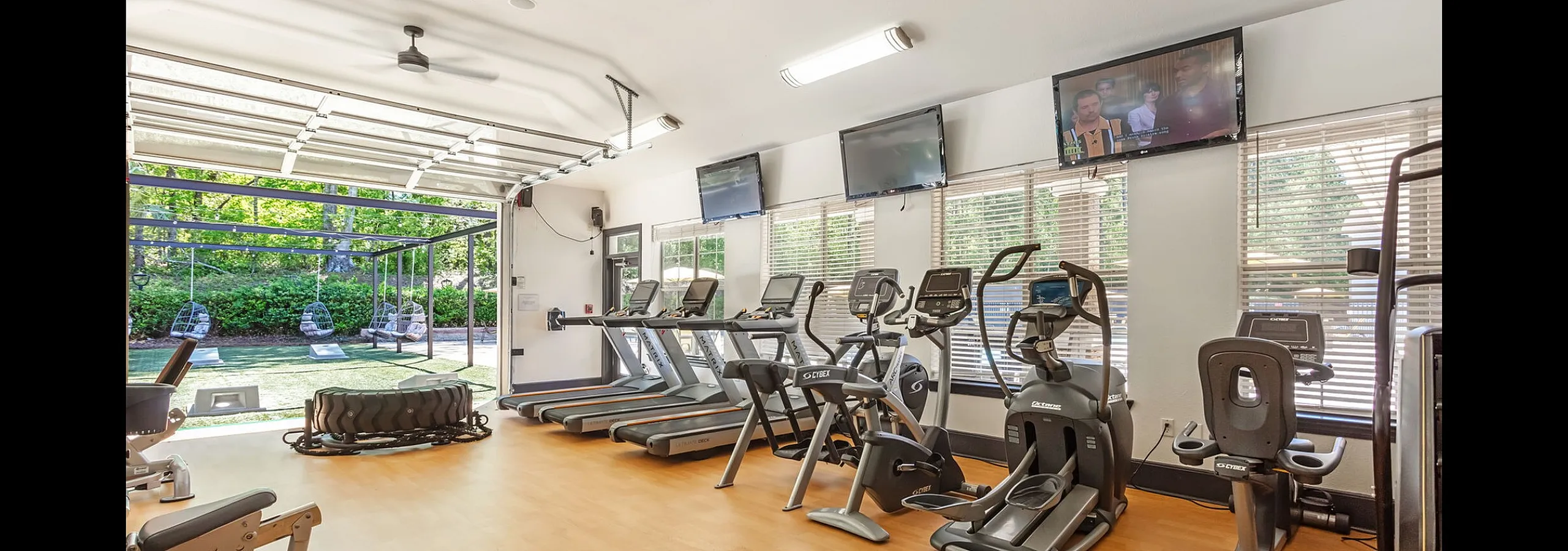 Modern fitness center with state-of-the-art gym equipment, including treadmills and elliptical machines, overlooking a lush garden with hanging chairs.
