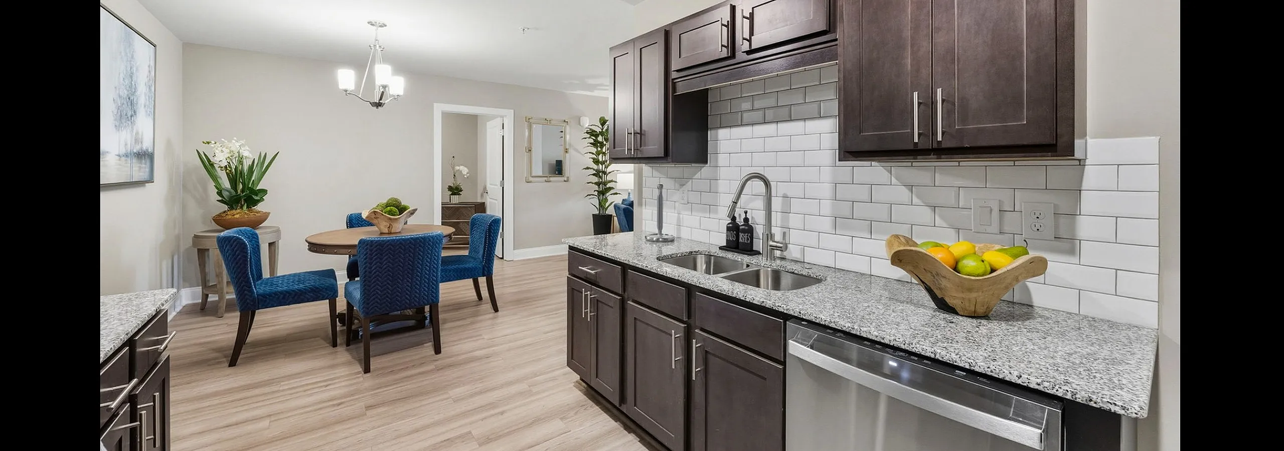 Contemporary kitchen and dining area with elegant blue chairs, sleek dark cabinetry, and stylish light fixtures, offering a warm and inviting ambiance.