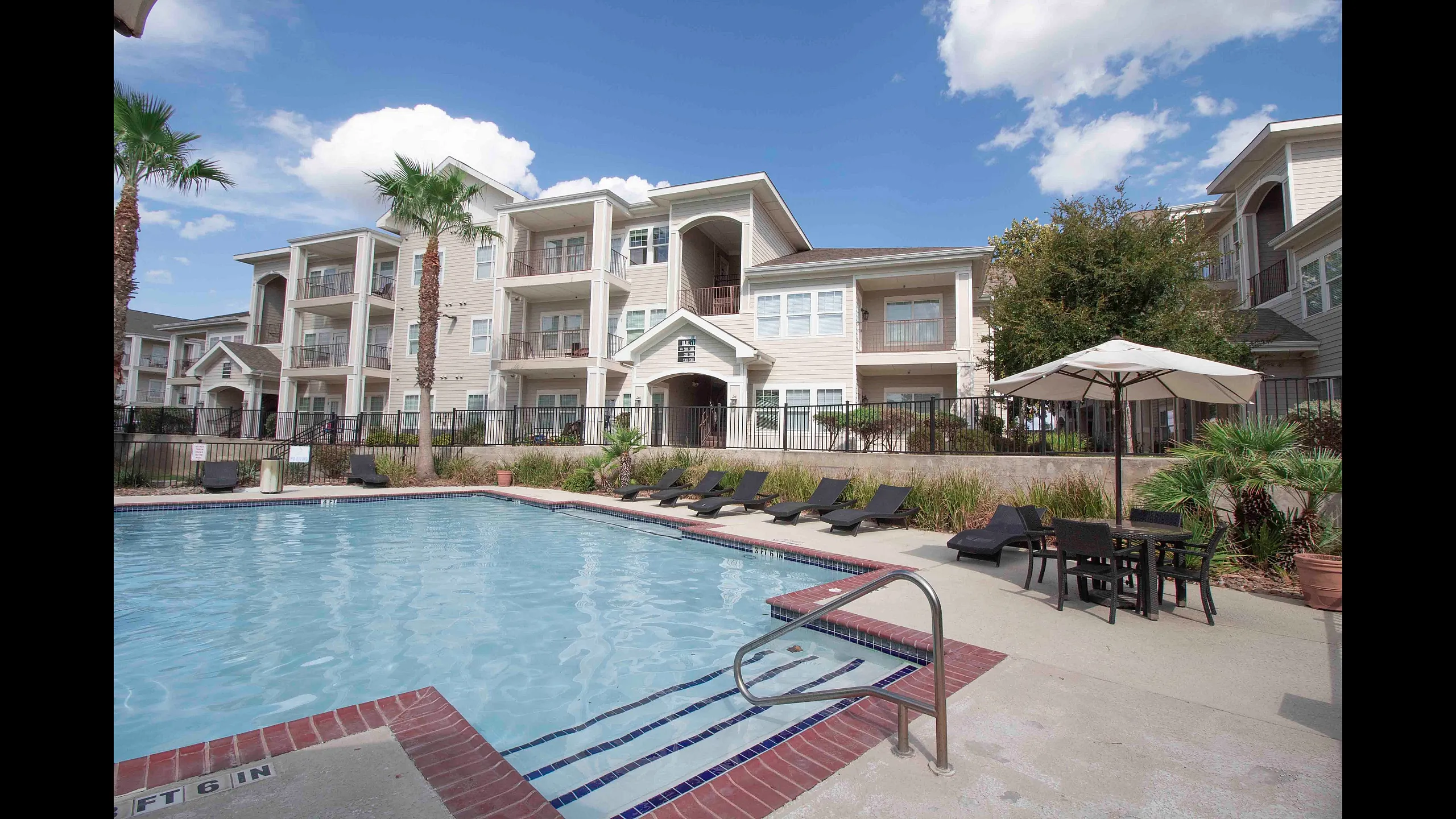 Inviting apartment complex exterior featuring a sparkling outdoor pool, elegant sun loungers, and vibrant landscaping beneath a clear blue sky.
