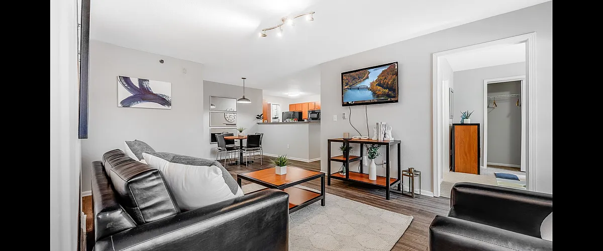 Modern open-plan living space featuring a cozy sitting area with sleek black leather couches, a dining area, and a well-appointed kitchen. Bright interior, stylish decor, and a mounted TV create a welcoming atmosphere.