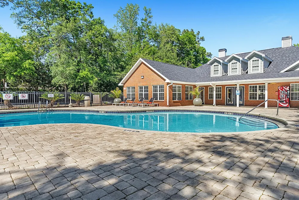 Inviting outdoor pool area surrounded by lush greenery, featuring a charming brick clubhouse with elegant lounge chairs and vibrant blue sky.