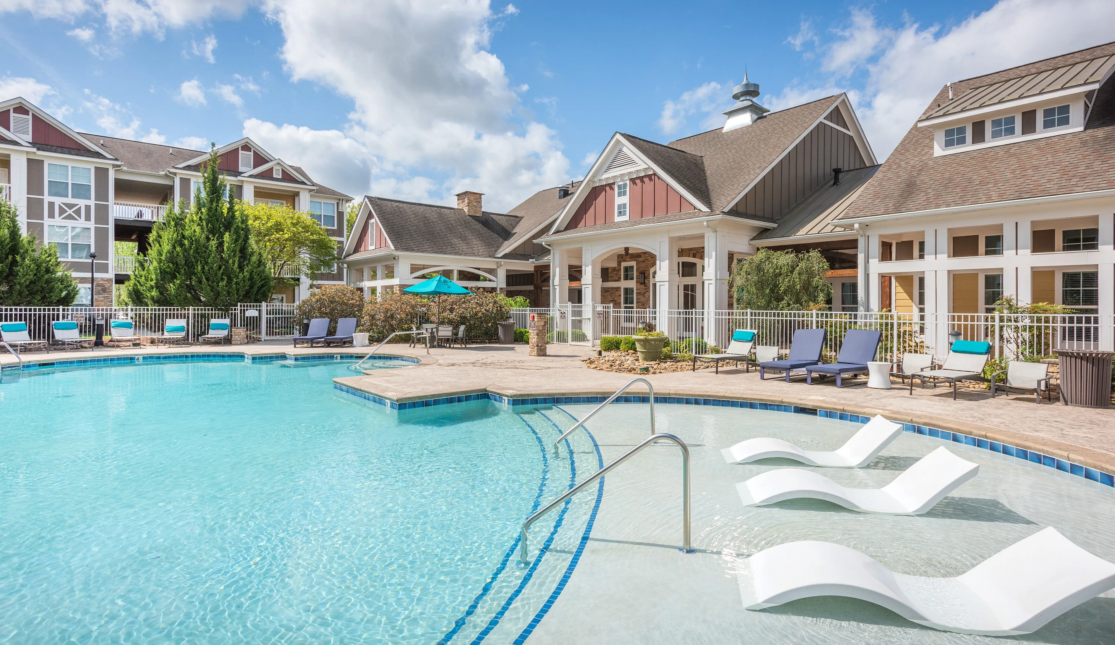 Charming apartment complex with a luxurious outdoor pool, surrounded by elegant lounge chairs and vibrant landscaping under a beautiful blue sky.