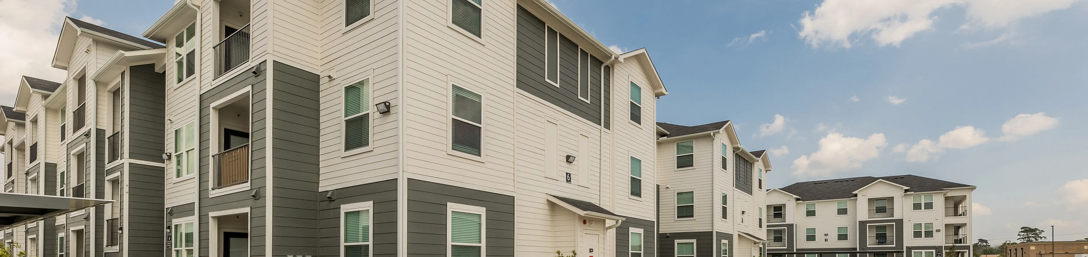 Charming modern apartment complex featuring multi-level buildings with balconies, surrounded by ample parking and manicured landscaping under a clear blue sky.