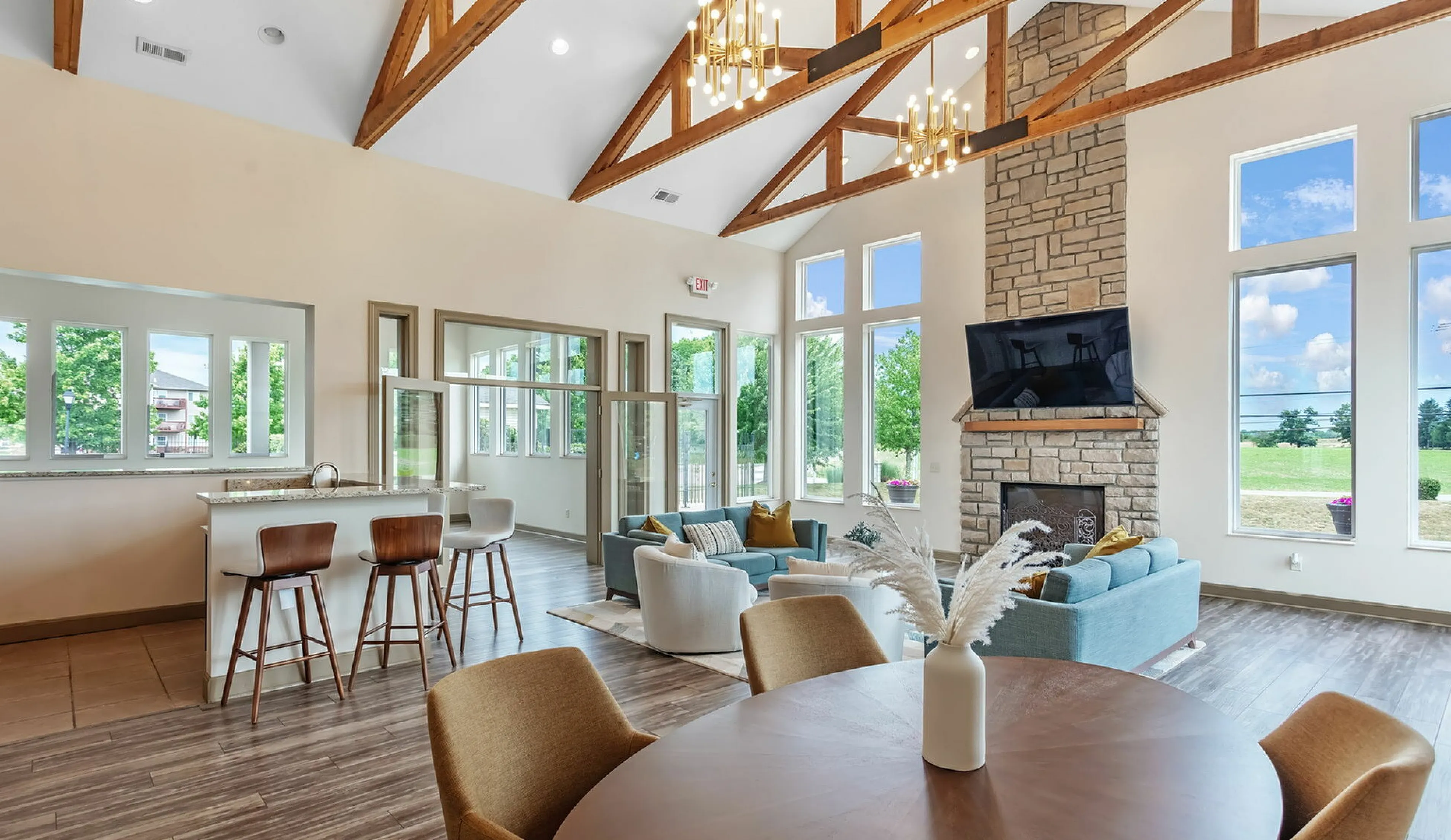 Bright and airy living space featuring vaulted ceilings, elegant wooden beams, and large windows with scenic views. Includes a stylish stone fireplace, cozy seating, and a modern open kitchen with bar stools.
