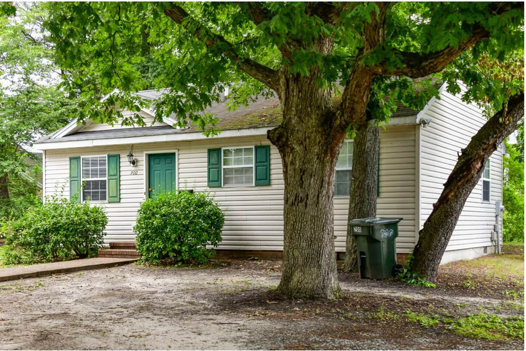 Charming single-story home surrounded by lush trees and greenery, featuring a quaint front porch with green shutters.