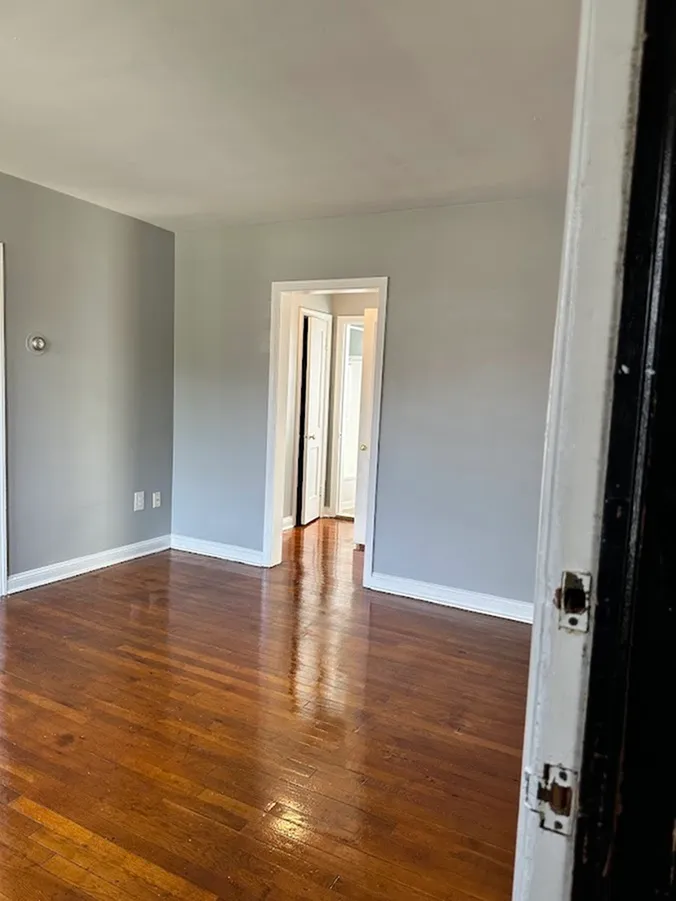 Bright room with polished hardwood floors and light gray walls, leading to a hallway with white doors.