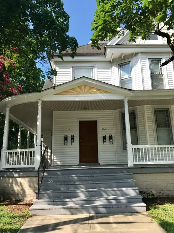 Charming Victorian-style house with a welcoming wraparound porch and elegant white exterior surrounded by lush greenery.