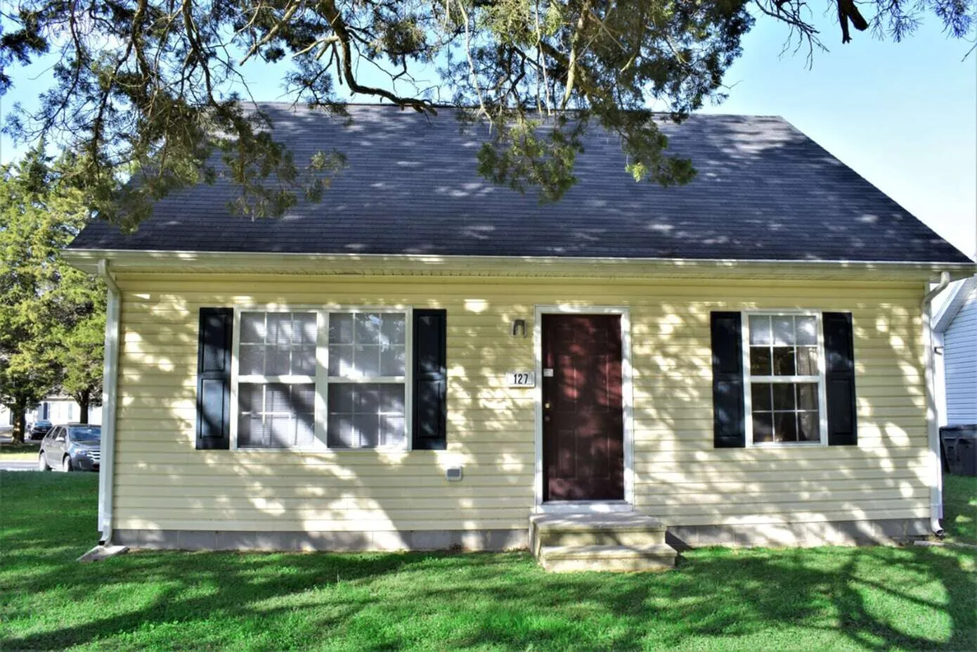 Charming yellow cottage with dark shutters and a welcoming front door, surrounded by lush green lawn and shaded by trees.